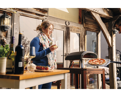 Scen med pizzaugn, pizza på en pizzaspade, bord med vinflaskor, glas och en kvinna med ett glas vin.