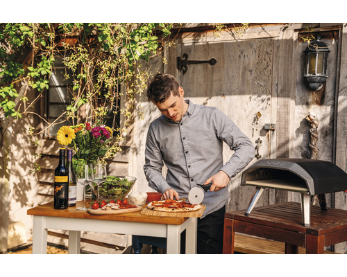 Man skär pizza i trädgården med pizzaugn, bord, vin, sallad och blommor.