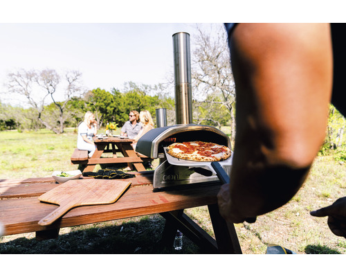 Utomhusscen med en pizzaugn, en pizza på en pizzaspade och en grupp människor vid ett picknickbord.