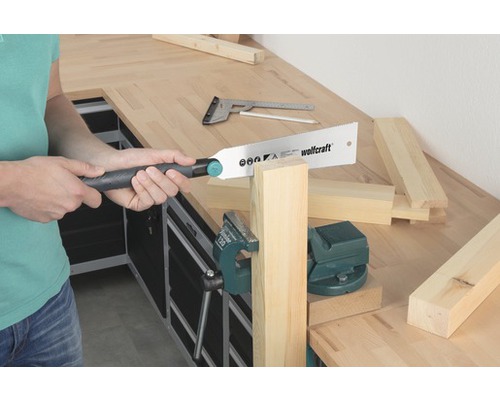 Person sågar trä med en handsåg vid en arbetsbänk. Skruvstycke håller fast arbetsstycket.