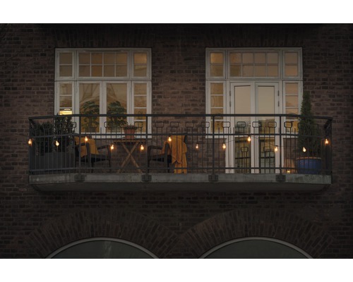 Dekorerad balkong med ljusslinga, stolar, bord och växter framför en tegelbyggnad.