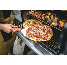 Person med pizzaspade framför grill med pizza, kycklingvingar och grönsaksspett
