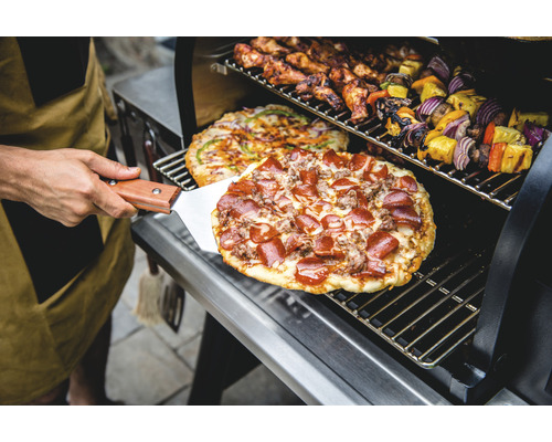 Person med pizzaspade framför grill med pizza, kycklingvingar och grönsaksspett