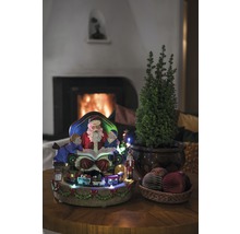 Juldekoration med jultomte, barn, tåg, litet träd och dekorativa bollar på ett bord framför en öppen spis.