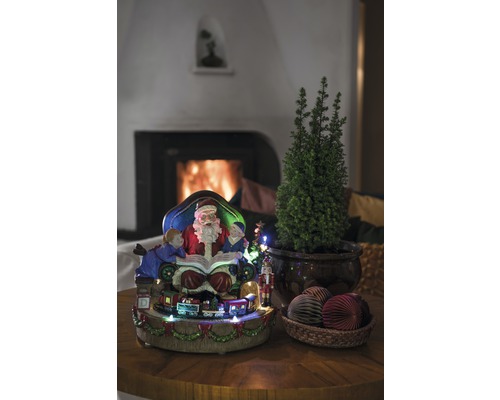 Juldekoration med jultomte, barn, tåg, litet träd och dekorativa bollar på ett bord framför en öppen spis.