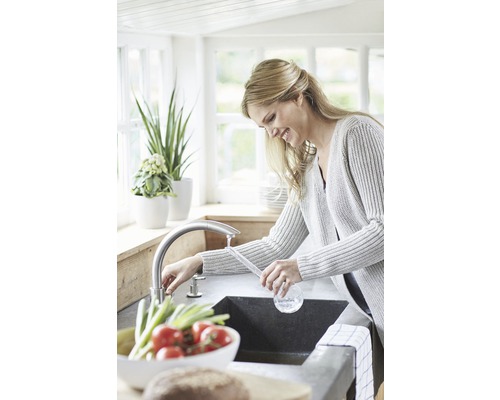 Kvinna vid diskbänken i ett ljust kök med armatur och färska grönsaker