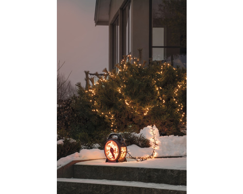 Dekorativt ljuslykta med fågelmotiv och ljusslinga på en snöig trappa framför ett hus.