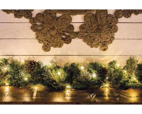 Dekorativ scen med jutekrans, grangrönska, kottar och ljusslinga på ett träbord framför en trävägg.