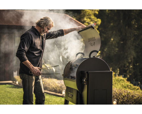 En man med grilltång öppnar locket på en grill utomhus, rök stiger upp.