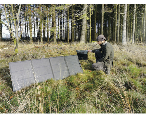 Man sitter på huk i skogen och använder en solcellsmodul för att ladda en generator