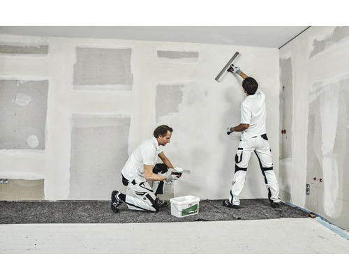 Två hantverkare spacklar en vägg i ett rum.