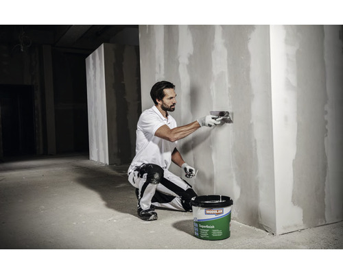 En man jämnar ut en vägg med spackel, medan en hink med spackel står på golvet.