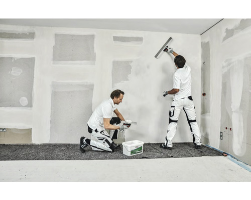 Två hantverkare spacklar en vägg inomhus.