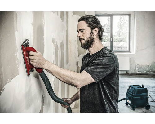 Hantverkare slipar vägg med en handslipmaskin ansluten till en dammsugare.