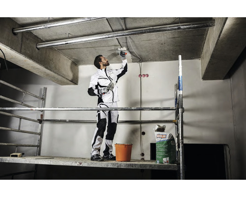 En hantverkare på en ställning spacklar ett tak. En säck med spackel och en hink står på ställningen.