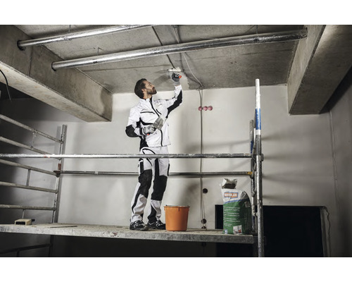 Hantverkare på en byggnadsställning som applicerar takputs med en spackel. I bakgrunden finns en säck med puts.