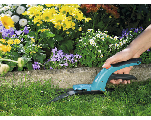 En person använder en Gardena grässax i trädgården för att klippa gräs, med blommor i bakgrunden.