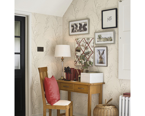 Hörnvy av ett rum med skrivbord, stol och dekorativa bilder på väggen, tapetserad med ett mönster av små blommor och kvistar.