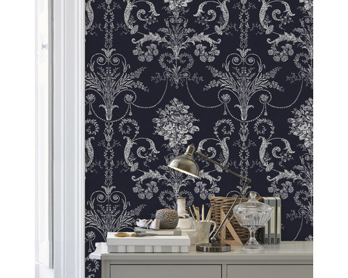 Närbild av ett rum med en vägg täckt av en mönstrad tapet. På ett bord står diverse dekorativa föremål och en lampa.