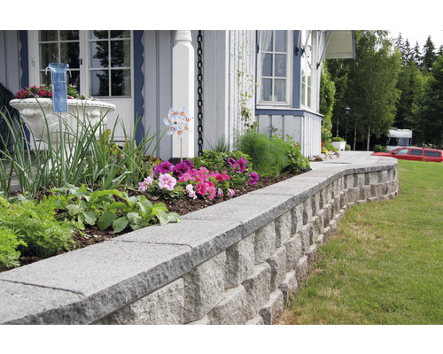 Trädgård med upphöjda blomrabatter och stenmur framför ett hus.