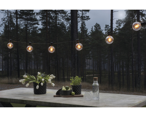 Scen med en ljusslinga, blommor och drycker på ett bord utomhus framför en skog