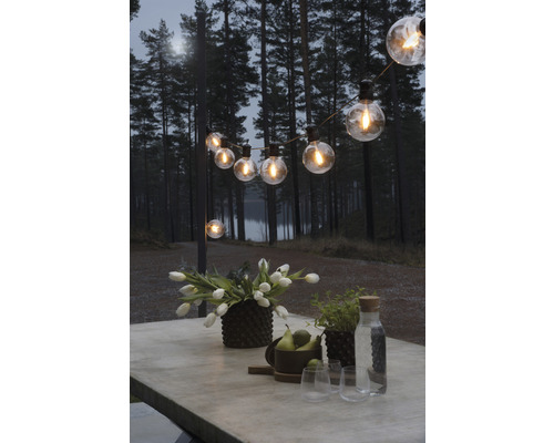 Dekorerat bord med blommor, frukt och glas under en ljusslinga utomhus.