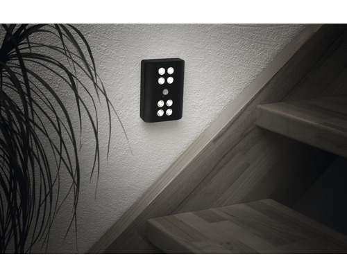 Svart vägglampa med rörelsedetektor bredvid en trappa och en växt