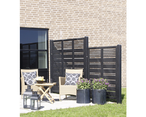 Terrass med vindskydd, rottingfåtöljer, bord, lyktor och blomkrukor mot en husvägg