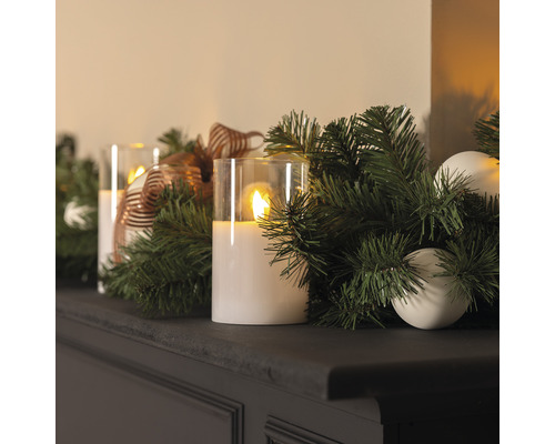 Dekorativt arrangemang med LED-ljus, grangrönt och ornament på en hylla.