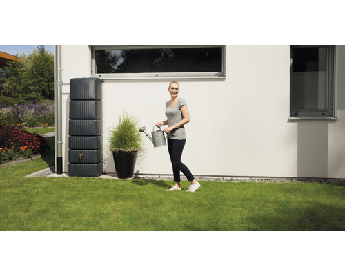 Scen med regnvattentank, kvinna med vattenkanna och växt i kruka i trädgården.