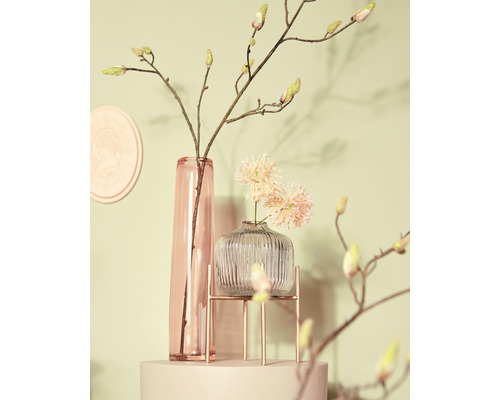 Dekorativ scen med en smal glasvas, en räfflad glasvas på ett metallstativ och grenar med knoppar.