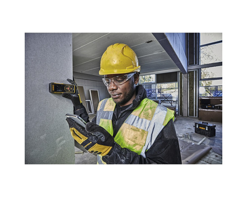 En byggnadsarbetare med skyddshjälm och skyddsglasögon använder en laseravståndsmätare på en vägg på en byggarbetsplats.