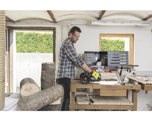 En man arbetar med en vedklyv i en verkstad. Vedträn ligger på bordet.