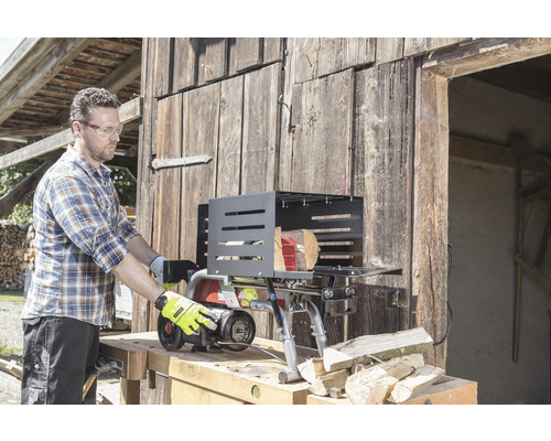 En man använder en vedklyv framför en trävägg. Vedklyven står på en arbetsbänk och det ligger vedträn bredvid honom.