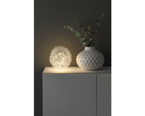 Dekorativ bordslampa i form av ett klot bredvid en vas med växter på ett skåp