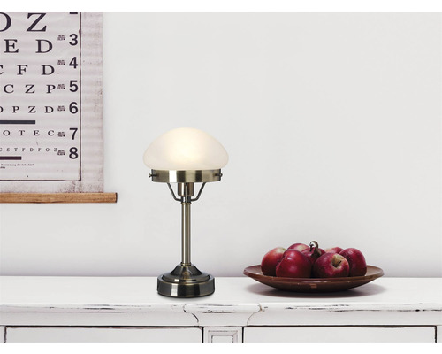Bordslampa med glasskärm och metallfot på ett bord med äpplen och en syntavla