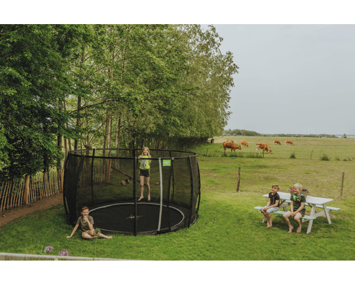 Trädgårdsscen med barn på en rund studsmatta med säkerhetsnät och en picknickbänk, med en äng med kor i bakgrunden