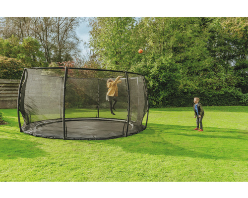 Rund trädgårdstrampolin med säkerhetsnät där ett barn leker med en boll medan ett annat barn tittar på.