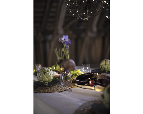 Dekorativt bord med kronärtskockor, glas, blomvas och ljusslingor under sfäriska lampor.