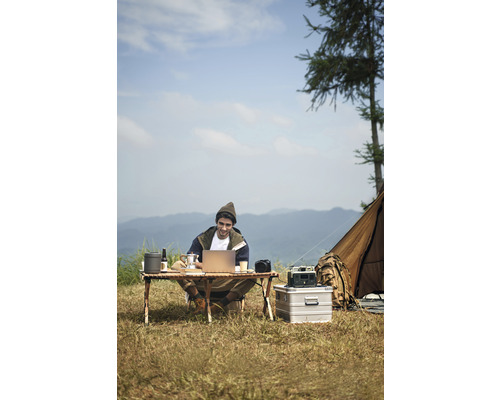 Man sitter vid ett campingbord och arbetar på en bärbar dator utomhus med ett tält i bakgrunden.