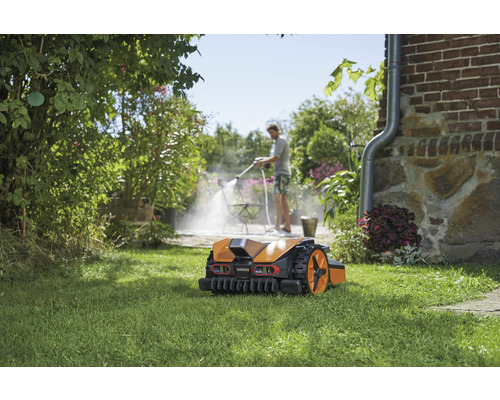 Robotgräsklippare på gräsmatta, i bakgrunden rengör en person en terrass med en högtryckstvätt