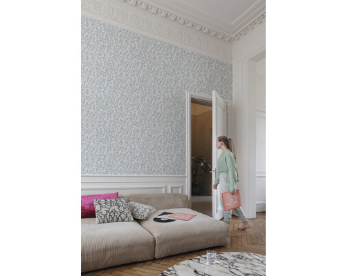 Ljust rum med blommönstrad tapet, soffa och en ung kvinna som går genom en öppen dörr