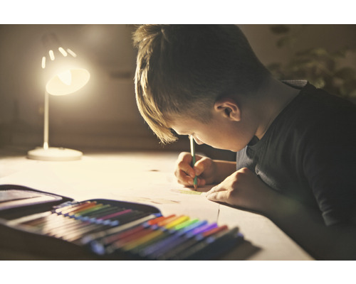 Pojke ritar med färgkritor under en lampa. Pennfodral med färgkritor på bordet.