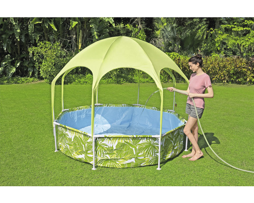 Trädgårdsscen med en ram бассейн med bladmönster och solskyddstak, medan en kvinna fyller poolen med en trädgårdsslang.