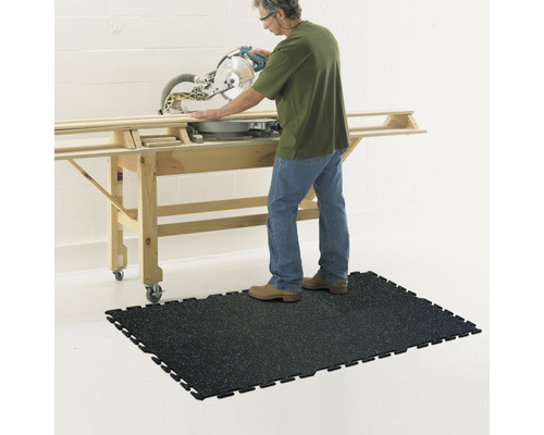En person använder en kap- och gersåg på ett arbetsbord, stående på en svart verkstadsmatta.