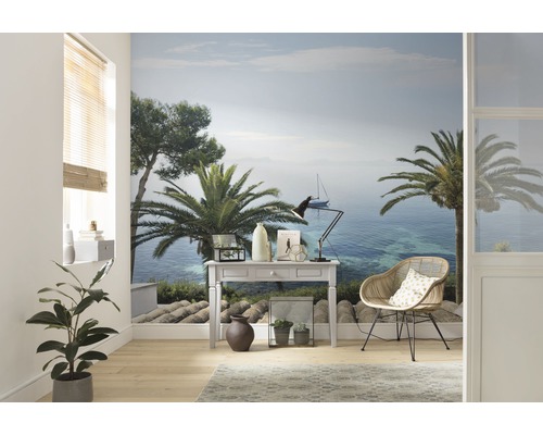 Vardagsrum med fototapet som visar ett kustlandskap med palmer och en segelbåt, inrett med ett skrivbord och en rottingfåtölj.