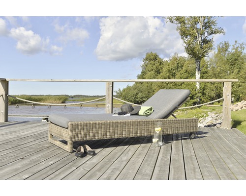 Flätad trädgårdssäng med grå klädsel, hatt, handduk och dryck på en träterrass vid sjön