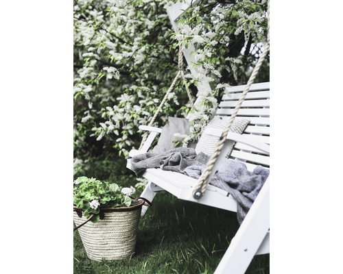 Trädgårdsscen med vit gunga, kuddar, filt, blommor och korg i trädgården