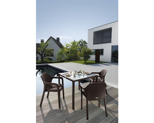 Trädgårdsscen med ett fyrkantigt trädgårdsbord och fyra matchande karmstolar på en träterrass intill en pool och ett modernt hus.