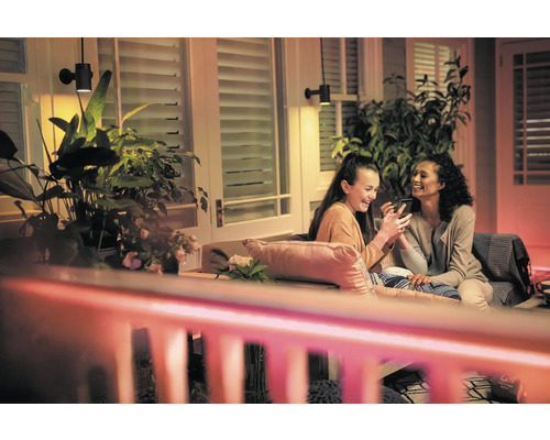 Scen med två kvinnor som sitter på en terrass under utomhusvägglampor och tittar på en smartphone, omgiven av växter.